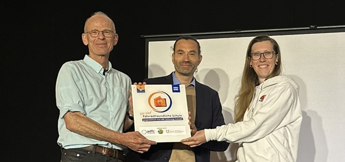 Nora Müller (links) vom ADFC SH übergibt die Plakette "Fahrradfreundliche Schule" an Mathias Kadow-Leder (Schulleiter Holstenschule) und Christian Wagner (Fahrradbeauftrager Holstenschule Neumünster)